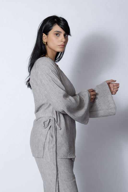 امرأة ترتدي طقماً رمادياً أنيقاً مكوناً من بلوزة برباط جانبي بأكمام طويلة وبنطال واسع الأرجل، مع حذاء كعب باللون الأزرق الفاتح، تقف بثقة أمام خلفية بيضاء وتضع يدها برقة على رقبتها