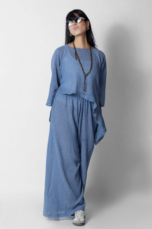 امرأة ترتدي طقماً واسع الأرجل من قماش الدنيم الأزرق الفاتح، يتكوّن من بلوزة بأكمام طويلة وبنطال واسع بخصر مطاطي، مع عقد طويل ونظارات شمسية، تقف بثقة وتضع يدها فوق رأسها، في خلفية بيضاء بسيطة